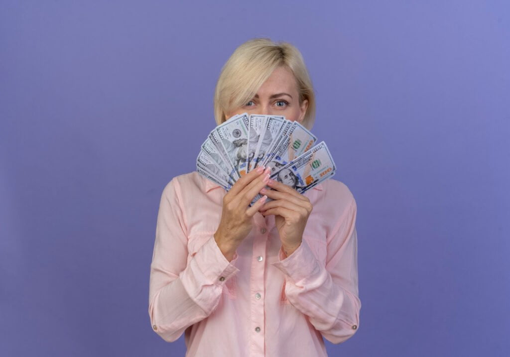 young blonde slavic woman holding money looking camera from money isolated purple background with copy space